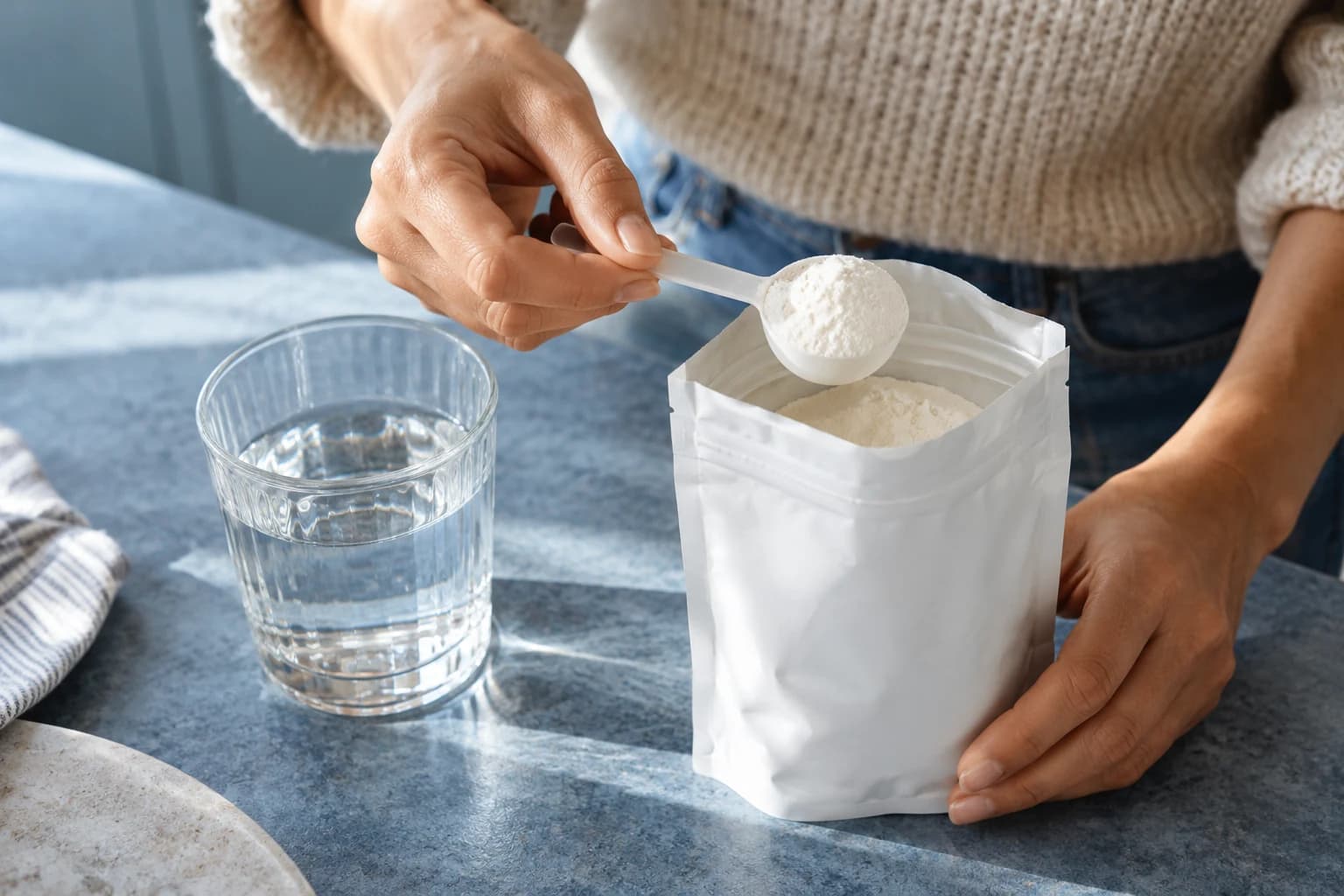 Hands holding an open white powder pouch and a filled scoop beside a glass of water.