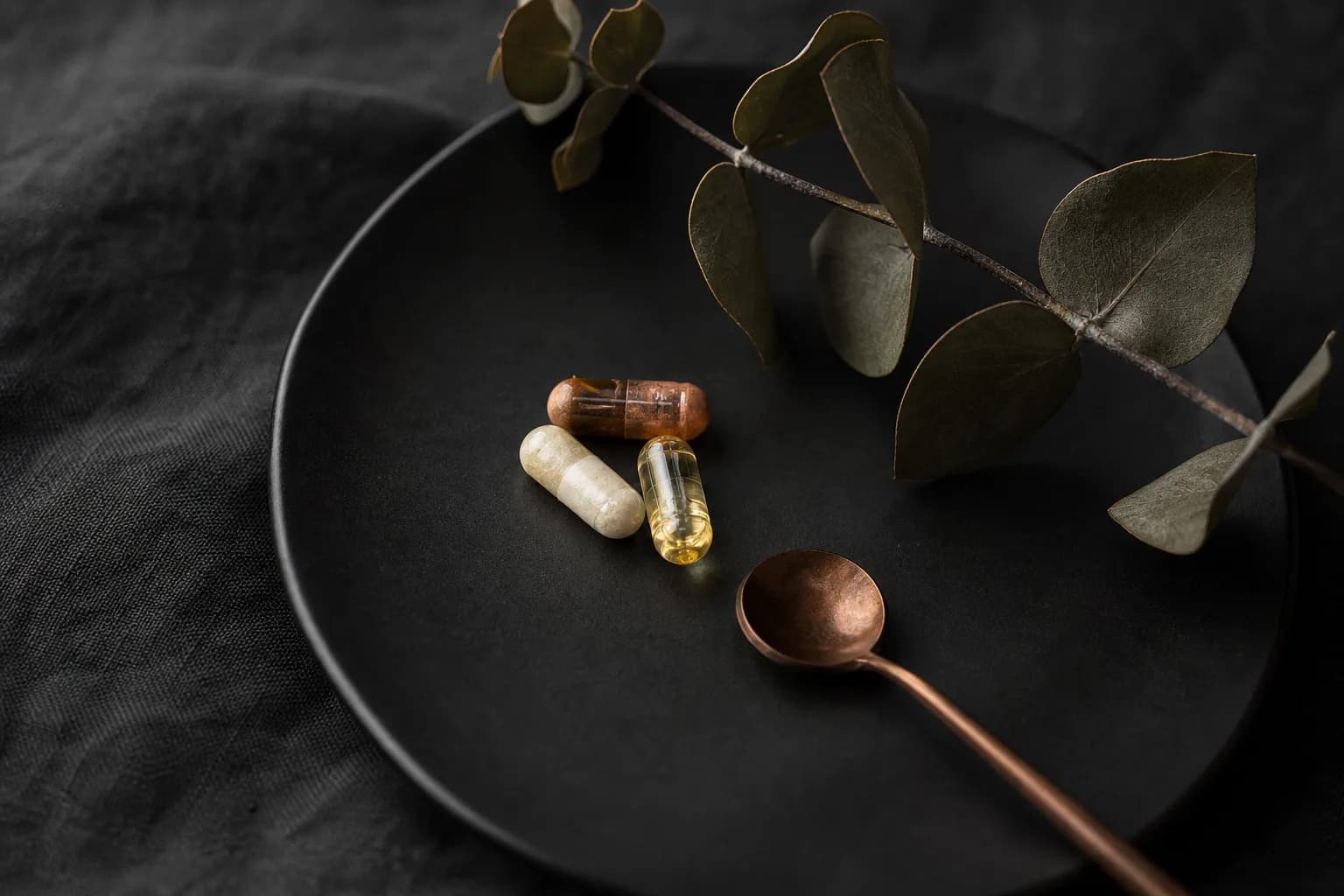 Three capsules and a small copper spoon on a dark plate beside a leafy branch.