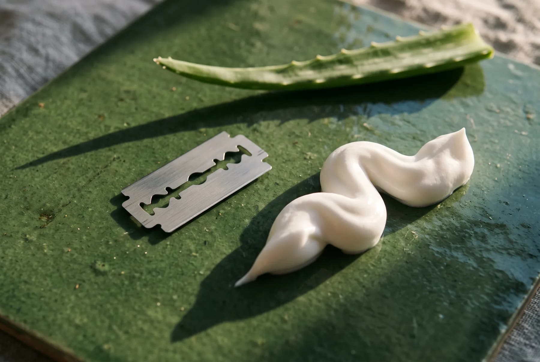 A fresh razor blade and a ribbon of shaving cream arranged with an aloe leaf
