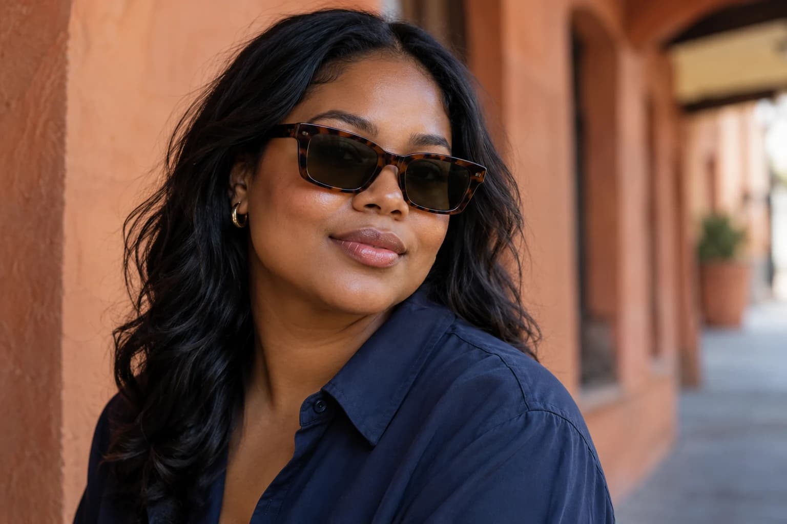 Woman wearing dark rectangular sunglasses beside a terracotta-colored wall.