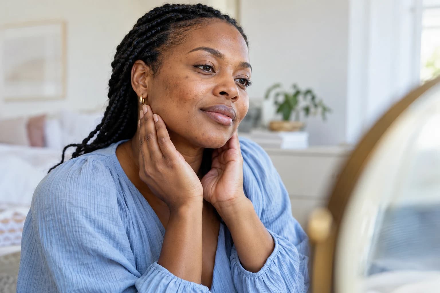 Woman with braided hair resting her hands along her jaw while looking toward a mirror.