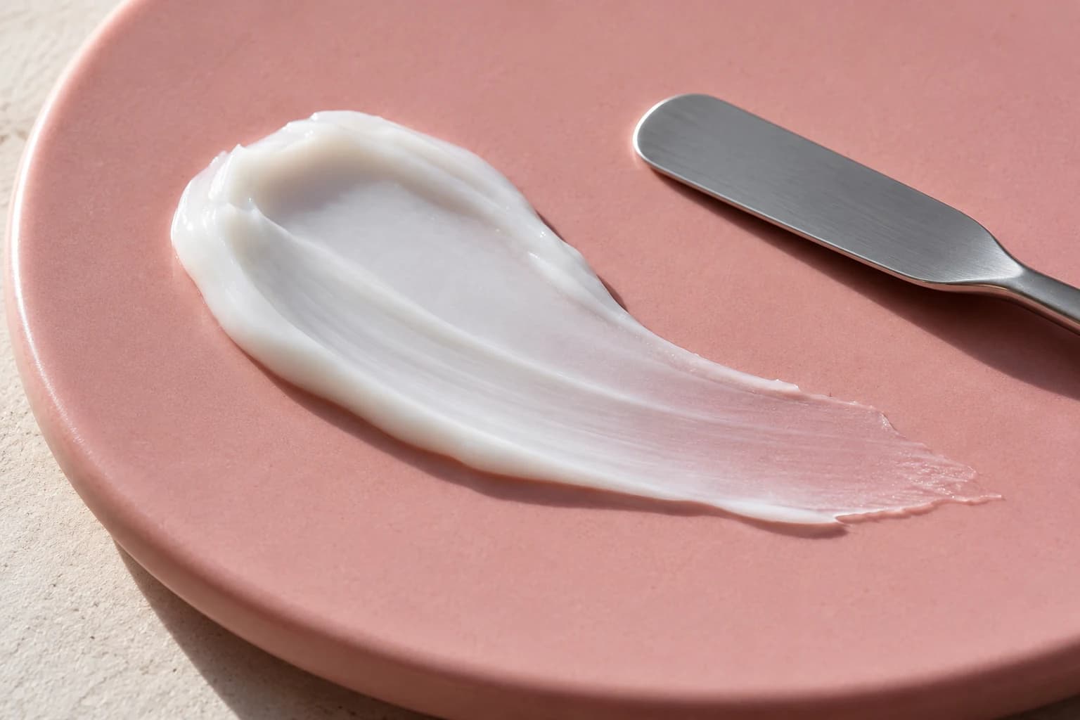 White cream swatch on a pink ceramic surface beside a metal spatula.