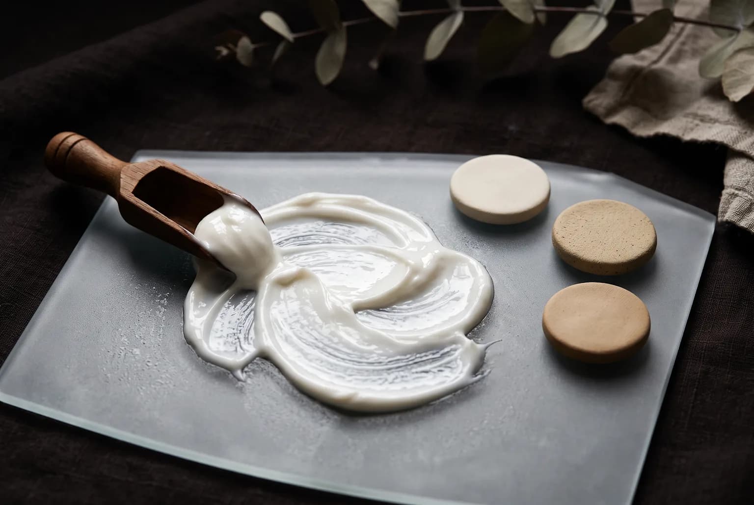 Rich moisturizer swirled across frosted glass beside neutral ceramic discs