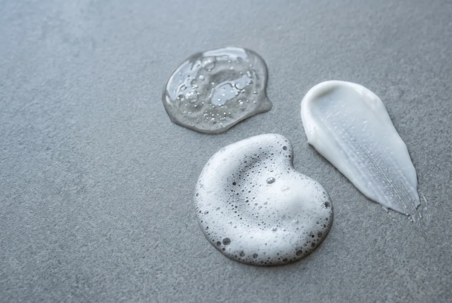 Close-up of three cleanser textures—gel, foam, cream—on a matte tile