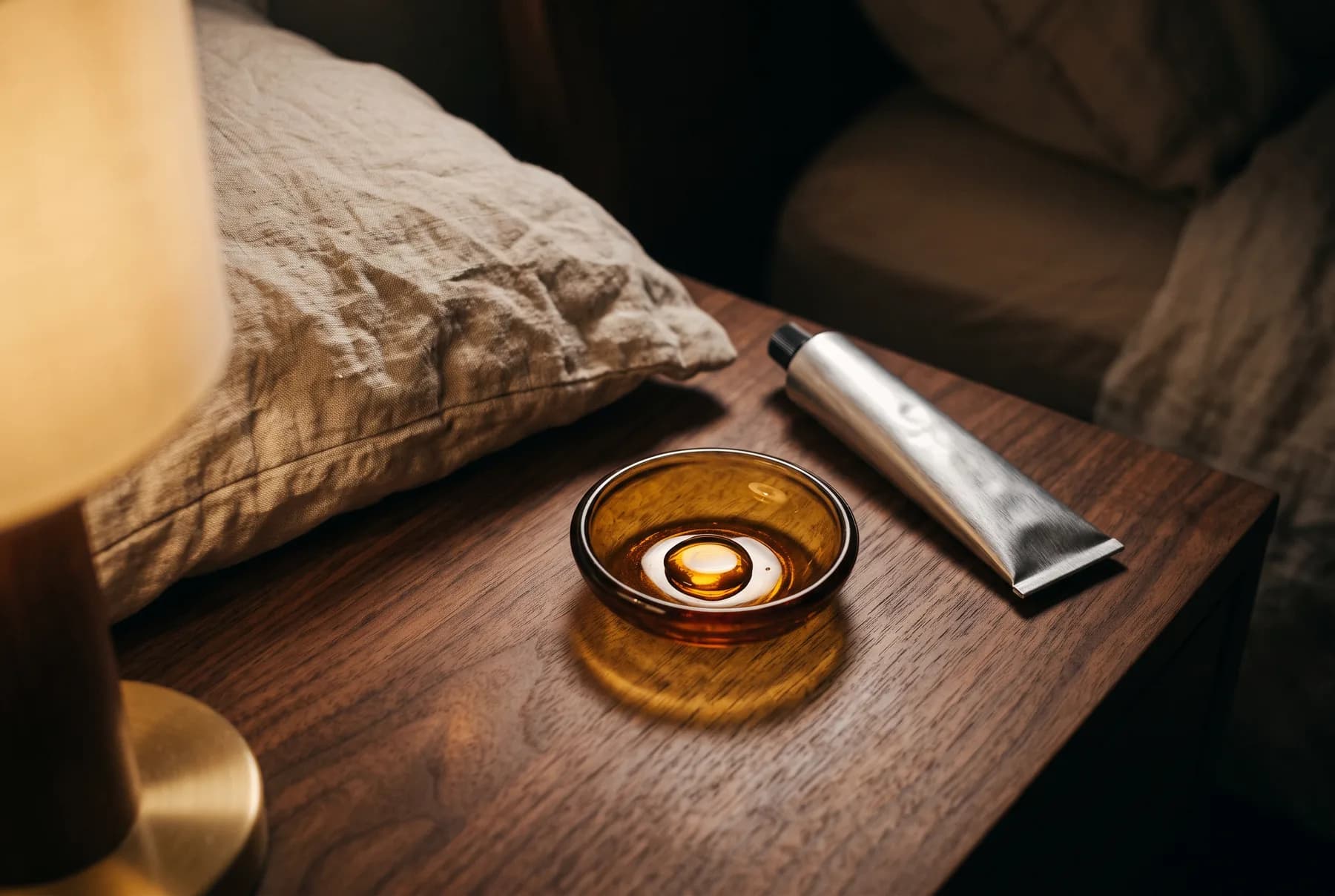 Nighttime skincare objects styled beside linen bedding under warm lamplight